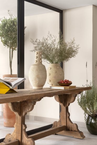 Rustic wooden table adorned with unique ceramic vases and greenery, showcasing an open book, set against a modern glass backdrop with reflections of potted plants