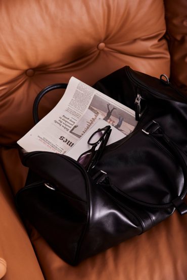 Black leather travel bag with a newspaper and sunglasses on a brown leather couch at White City Limassol Hotel.